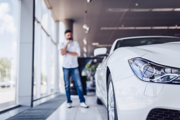 Vue de face d'une voiture blanche dans un salon de concession avec un homme pensif sur le plan flou d'arrière-plan