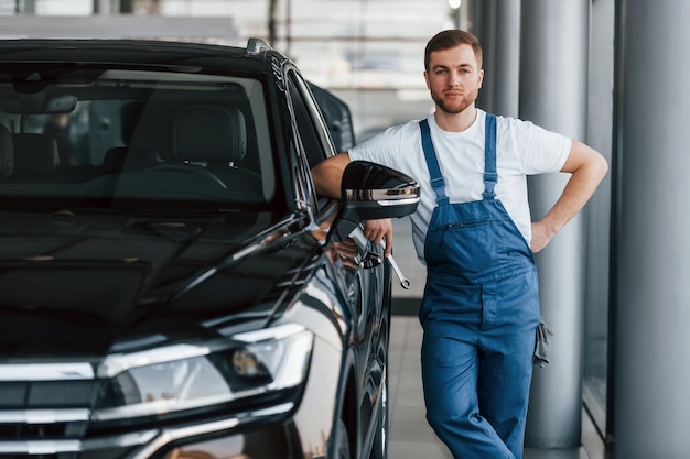 Vue de face de la voiture Jeune homme en chemise blanche et uniforme bleu répare l'automobile