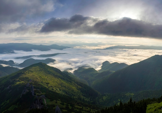 Vue fantastique sur la vallée de montagne recouverte de bas bouffants blancs comme des nuages de neige s'étendant jusqu'à l'horizon brumeux sous un ciel matinal lumineux avec une lueur orange clair au lever du soleil. Beauté du concept de la nature.