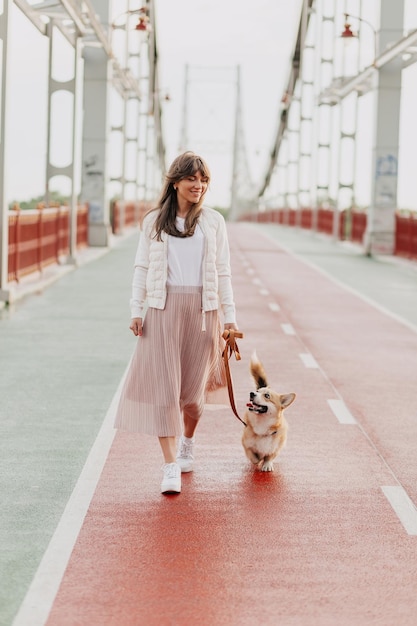 Vue d'une femme européenne aux longs cheveux lâches portant une longue jupe rose et une veste blanche marche avec son chien sur le pont de la ville le matin d'été