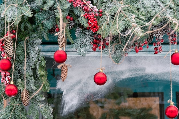 Vue d'une fenêtre enneigée décorée de branches de sapin, de fruits rouges et de boules, d'une corde, de pommes de pin et de pommes de pin.