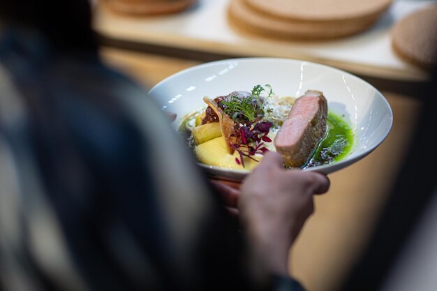 Vue de fond d'une personne tenant une assiette de croûte d'agneau rôtie servie avec de la sauce et des légumes verts