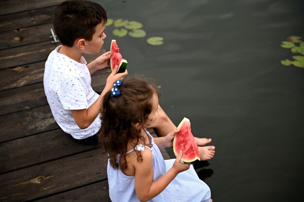 Vue en grand angle de deux enfants mignons, garçon et fille, avec des tranches de pastèque dans les mains, assis sur la jetée avec leurs jambes abaissées dans l'eau et profitant des soirées d'été à la campagne.