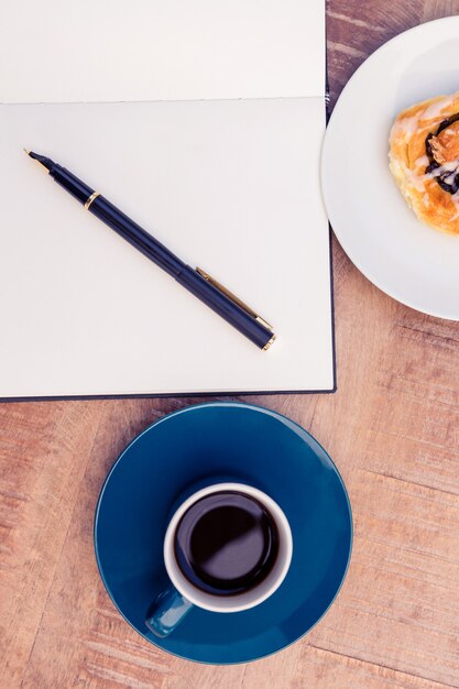 Vue grand angle du stylo sur le bloc-notes par la nourriture et le café à la table dans le bureau