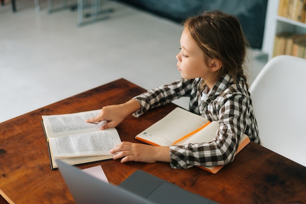 Vue en grand angle d'une écolière mignonne faisant ses devoirs en lisant un livre de papier assis à table dans une salle d'enfants claire. Vue de dessus d'une écolière du primaire étudiant à la maison, mise au point sélective.