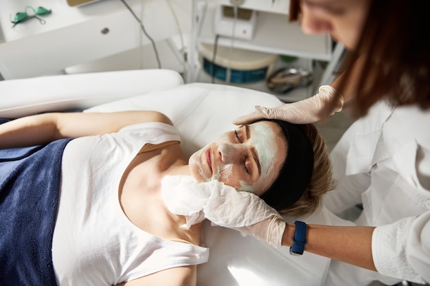 Vue en grand angle d'une esthéticienne effectuant une procédure de beauté sur le visage d'une femme, enlevant le reste du produit cosmétique à l'aide de serviettes humides spéciales. Soins de la peau professionnels dans une clinique de cosmétologie