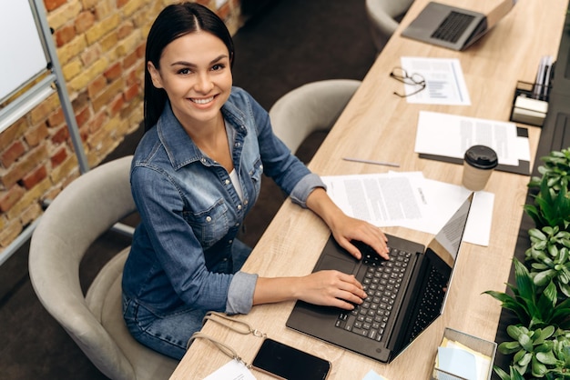 Vue grand angle de la femme brune assise à la table et tapant au clavier tout en travaillant dans son bureau avec plaisir sourire