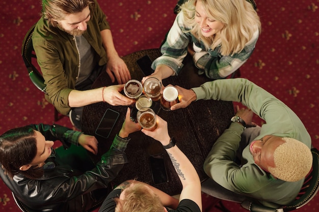 Vue grand angle d'un groupe d'amis assis à table en train de griller avec de la bière lors de leur réunion au bar