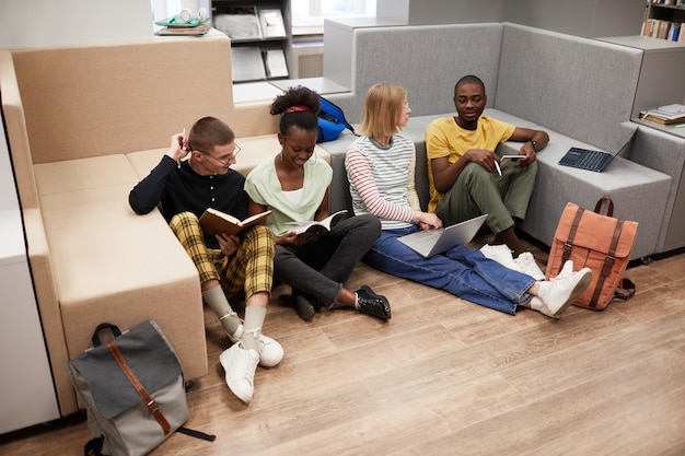 Vue en grand angle sur un groupe diversifié de jeunes étudiants qui étudient ensemble tout en étant assis par terre dans la colle...