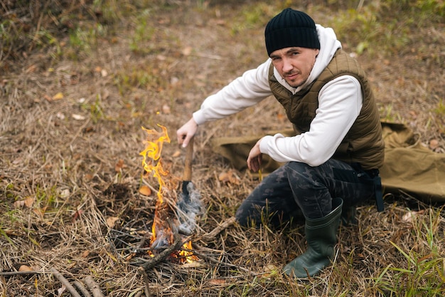 Vue en grand angle d'un homme voyageur confiant portant des vêtements chauds assis près d'un feu de camp brûlant et tenant une petite pelle regardant la caméra