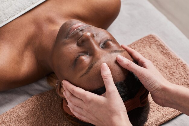 Vue grand angle sur une jeune femme afro-américaine profitant d'un massage du visage et souriant avec bonheur, avec des mains féminines en prise de vue