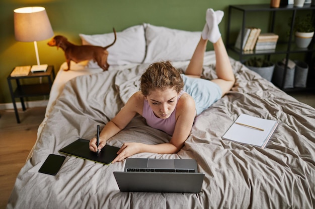 Vue grand angle d'une jeune femme dessinant sur une tablette graphique en position couchée dans son lit à la maison