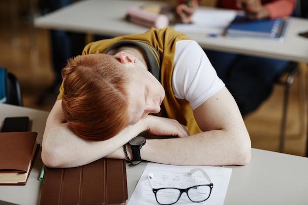 Vue grand angle sur un jeune homme aux cheveux roux endormi sur un bureau à l'école ou au collège