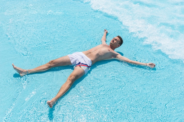 Vue grand angle d'un jeune homme portant un maillot de bain allongé sur le dos avec les bras et les jambes écartés dans les vagues peu profondes de la piscine par une journée ensoleillée en été