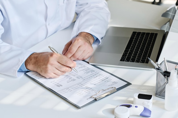 Vue grand angle d'un médecin mature remplissant la carte médicale du patient à table avec un ordinateur portable