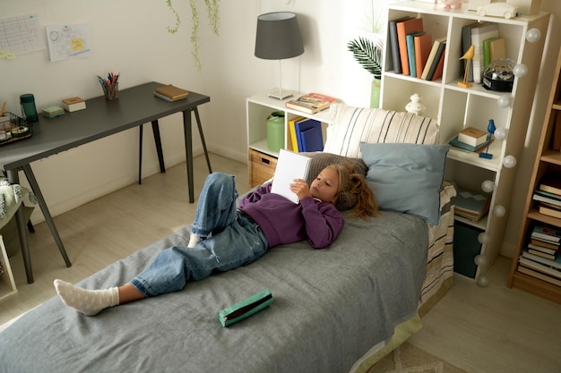 Vue grand angle de la petite fille allongée sur son lit et lisant son journal pendant son temps libre à la maison