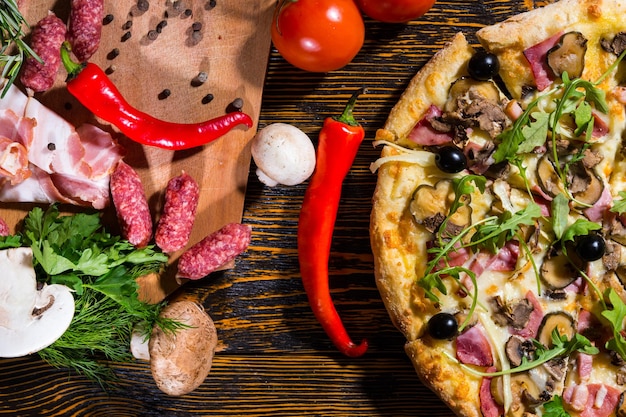 Vue grand angle de la pizza avec de la roquette et des olives sur une table en bois, près des tomates, des champignons et de la planche à découper avec du bacon