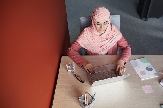 Vue de haut en bas à la jeune femme d'affaires portant un foulard à l'aide d'un ordinateur portable au bureau tout en travaillant au bureau, copiez l'espace