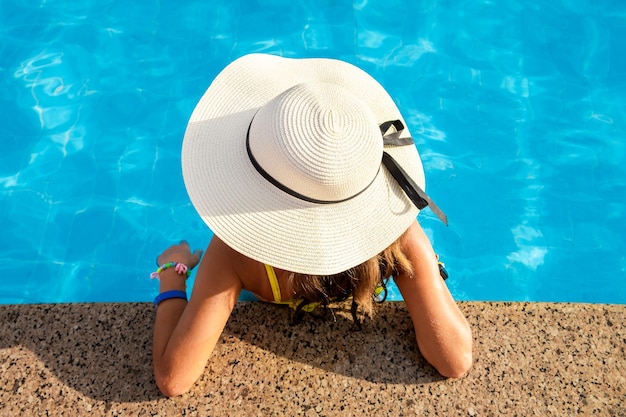 Vue de haut en bas d'une jeune femme portant un chapeau de paille jaune se reposant dans une piscine avec de l'eau bleu clair le jour ensoleillé d'été.