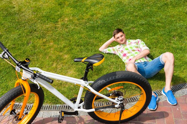 Vue de haut en bas sur un jeune homme relaxant allongé sur l'herbe à côté d'un vélo à pneus gras jaune et blanc