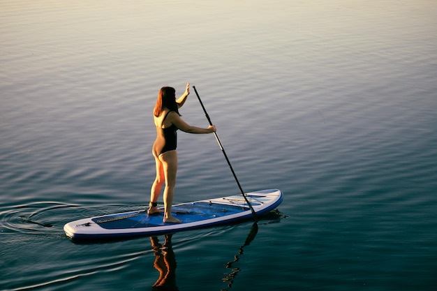 Vue d'en haut sur une femme d'âge moyen détendue sur une planche sup ramant avec une rame sur l'eau calme du lac Mode de vie actif Activité de loisirs pour les personnes d'âges différents