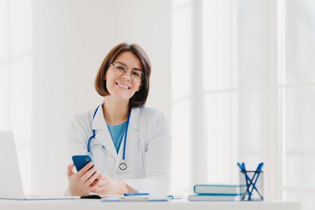 Vue horizontale d'un médecin professionnel souriant travaille dans des poses de clinique au bureau de l'hôpital moderne avec des gadgets électroniques envoie des messages texte sur le téléphone portable étant au travail Concept de technologie de soins de santé