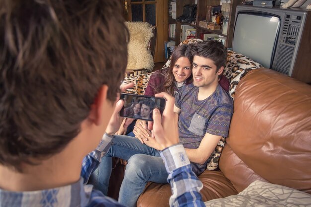 Vue d'un jeune ami prenant des photos avec un smartphone pour un couple d'adolescents hipster assis dans le canapé