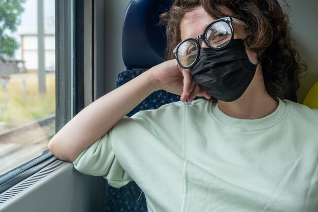 Vue d'une jeune femme voyageant en train pendant une pandémie portant des lunettes et un masque noir.