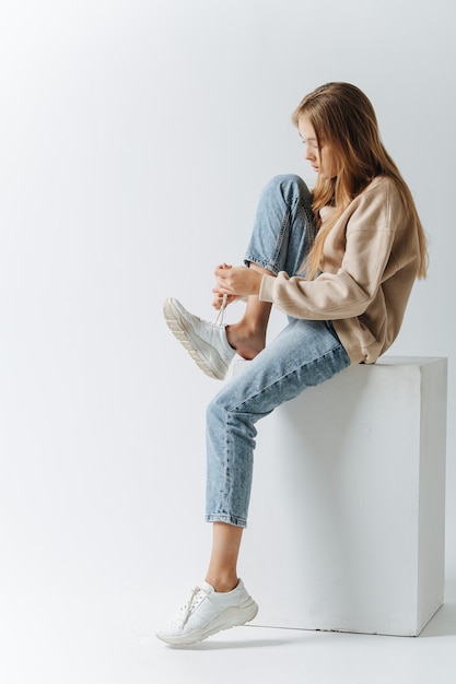 Vue latérale d'une adolescente laçant ses baskets. Assis sur un cube blanc. Dans un studio sur fond blanc.