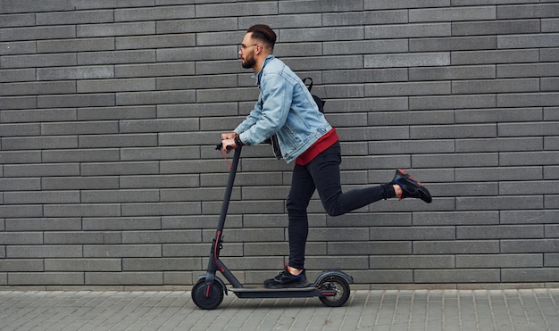 Vue latérale d'un beau jeune homme en vêtements décontractés qui fait du scooter électrique à l'extérieur pendant la journée ensoleillée