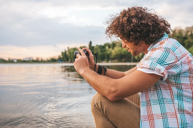 Vue latérale d'un beau mâle aux cheveux bouclés vérifiant des photos de la nature sur son appareil photo numérique Jeune bel homme porte une chemise décontractée avec un appareil photo numérique posant dans le parc