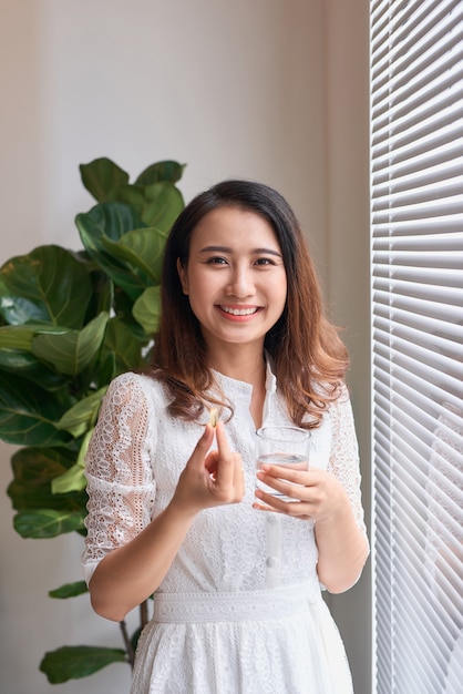 Vue latérale d'une belle femme prenant une capsule de vitamine avec un verre d'eau isolé en blanc