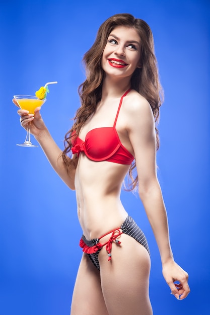 Vue latérale de la belle jeune brune en bikini rouge et bleu vif avec cocktail exotique souriant dos. Isoler.