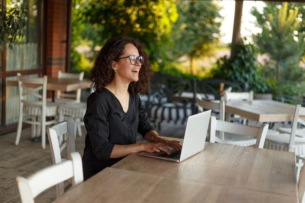 Vue latérale d'une belle jeune femme aux cheveux bouclés assis à la table en tapant sur l'ordinateur portable à la terrasse
