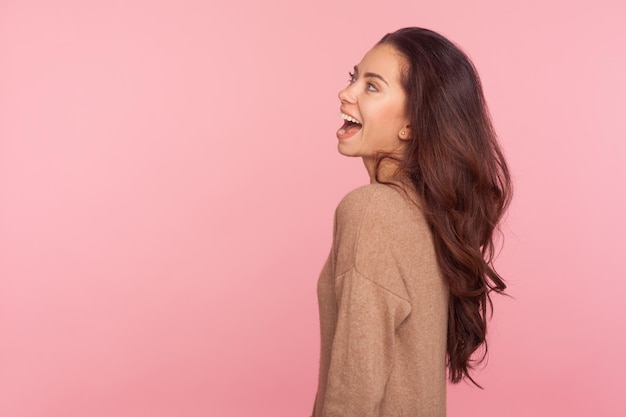 Vue latérale d'une belle jeune femme mince ravie avec de longs cheveux ondulés brune regardant avec un sourire joyeux extrêmement heureux exprimant l'optimisme des émotions positives studio intérieur tourné fond rose