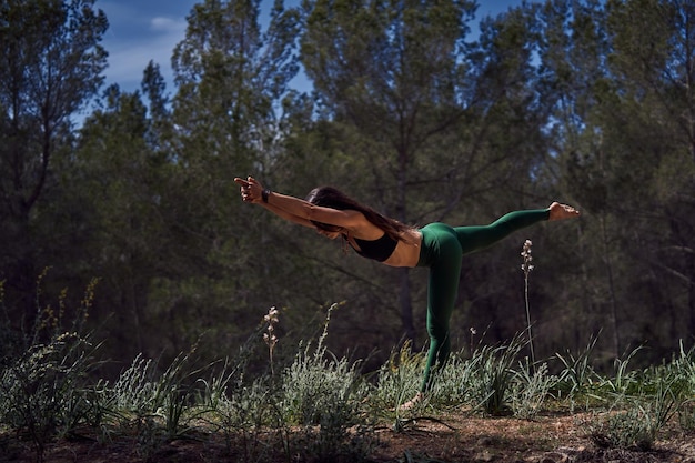 Vue latérale complète du corps d'une femme active pieds nus méconnaissable en vêtements de sport pratiquant la posture Virabhadrasana C pendant une séance de yoga en forêt