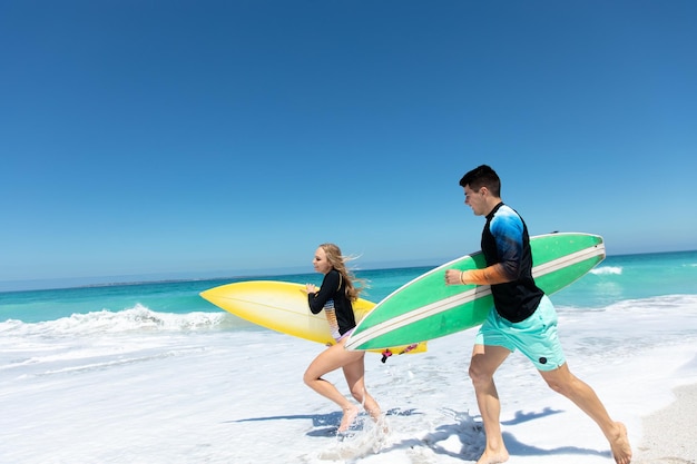 Vue latérale d'un couple caucasien courant sur la plage avec le ciel bleu et la mer en arrière-plan, tenant leurs planches de surf