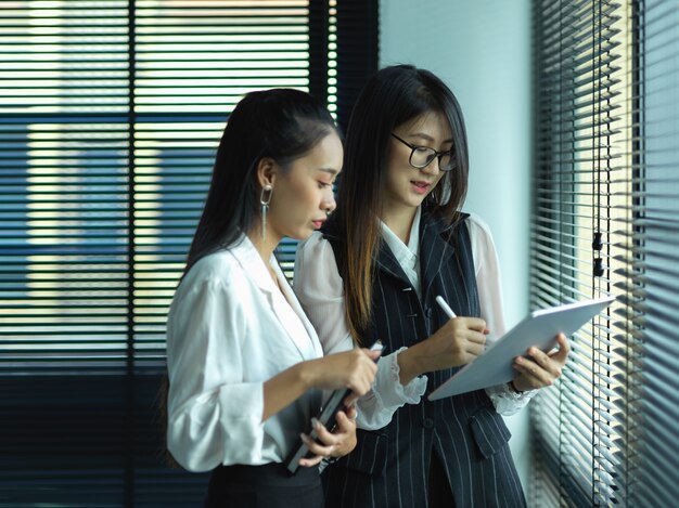 Vue latérale de deux femmes d'affaires consultant sur leur travail en se tenant debout à côté de la fenêtre au bureau