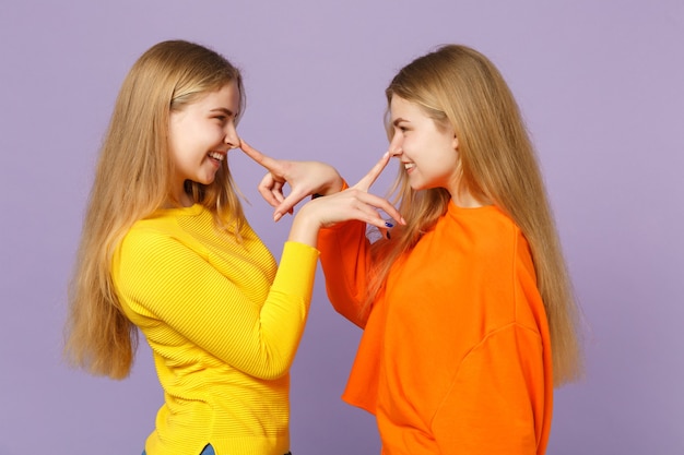 Vue latérale de deux filles jumelles blondes drôles dans des vêtements colorés mettant les doigts sur le nez s'amusant isolés sur un mur bleu violet pastel. Concept de mode de vie familial de personnes.