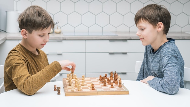 Vue latérale de deux frères amis préadolescents concentrés assis à une table blanche jouant aux échecs sur une planche de bois en compétition