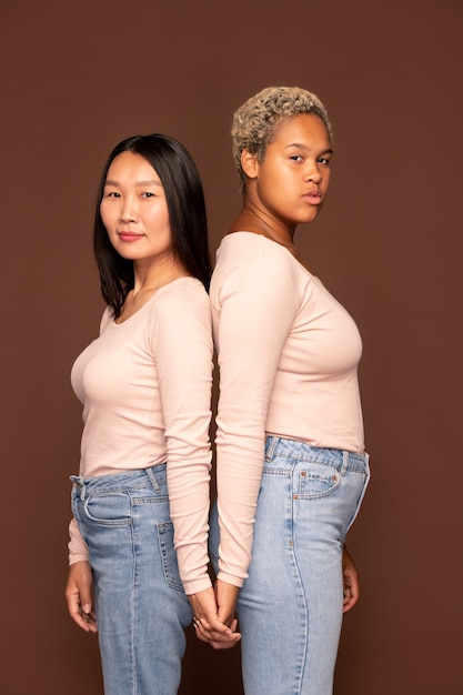 Vue latérale de deux jeunes femmes interculturelles en pulls blancs vous regardant tout en se tenant près l'une de l'autre sur fond marron