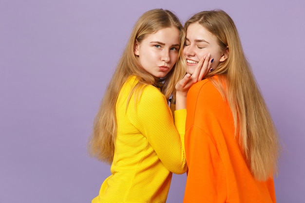 Vue latérale de deux tendres jeunes filles jumelles blondes mignonnes dans des vêtements colorés vifs regardant une caméra isolée sur un mur bleu violet pastel. Concept de mode de vie familial de personnes.