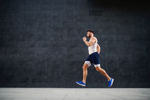 Vue latérale du bel homme musclé caucasien fit courir vite à l'extérieur. En arrière-plan, un mur gris.