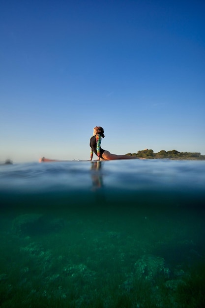 Vue latérale du corps entier d'une femme anonyme en maillot de bain faisant du Bhujangasana tout en pratiquant le yoga sur paddleboard dans la mer avec de l'eau turquoise claire sous un ciel du soir sans nuages