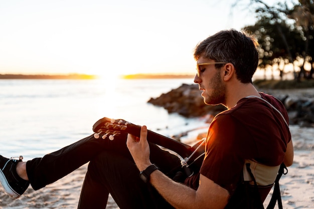 Vue latérale du jeune homme jouant de la guitare à l'extérieur avec bord de mer comme arrière-plan au coucher du soleil