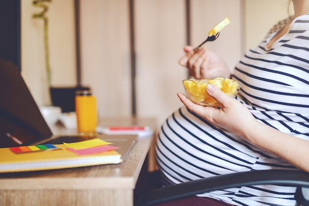 Vue latérale du jeune pigiste enceinte caucasien, manger des fruits et assis au bureau à domicile. Sur ordinateur portable de bureau, verre avec jus d'orange frais et cahiers. Concept de travail des femmes enceintes.