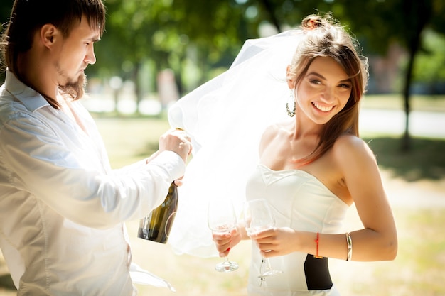 Vue latérale du marié barbu ouvrant une bouteille de champagne tout en souriant mariée tenant des lunettes. Dame charmante et bel homme célébrant le mariage nouvellement créé à l'extérieur. Notion de relations.