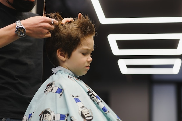 Vue latérale du mignon petit garçon se faire couper les cheveux par le coiffeur au salon de coiffure