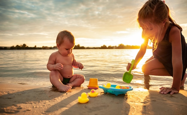 Vue latérale du petit bébé adorable jouant avec de minuscules canards jaunes en caoutchouc dans une petite piscine bleue et assis avec sœur aînée sur le sable de la plage