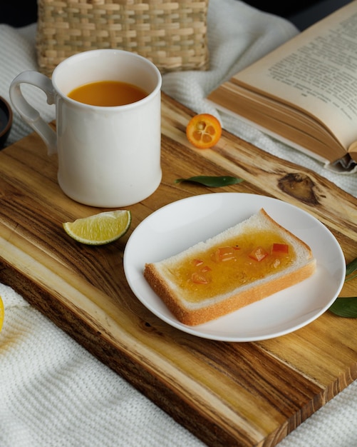 Vue latérale du petit-déjeuner avec une tranche de pain enduite de confiture dans une assiette et une tasse de jus d'orange tranche de citron vert avec des feuilles sur une planche à découper et un livre ouvert avec panier sur fond de tissu blanc